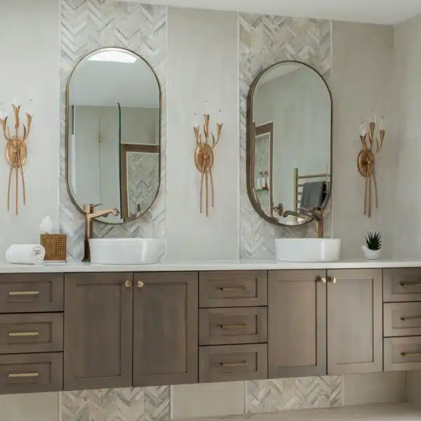 Bathroom renovation with floating midtone wood double vanity backdropped with a chevron tile detail, light color quartz counters, vessel sinks and tall brushed gold vessel faucets, stunning crystal wall sconces and unique oval mirrors in Medford, NJ.
