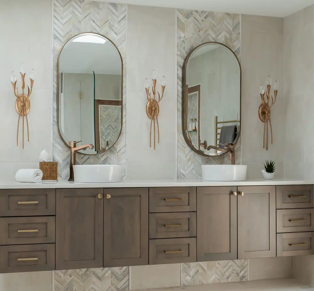 Bathroom renovation with floating midtone wood double vanity backdropped with a chevron tile detail, light color quartz counters, vessel sinks and tall brushed gold vessel faucets, stunning crystal wall sconces and unique oval mirrors in Medford, NJ.
