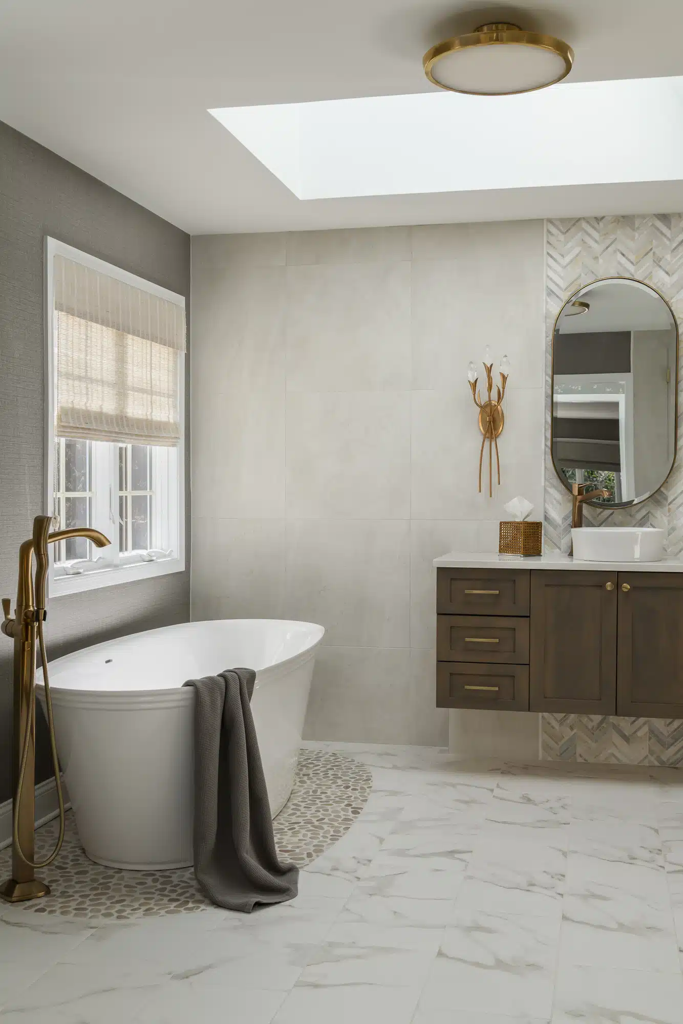 Freestanding deep oval tub with pebble floor accent underneath,champage bronze Brizo bath faucets, floating midtone color vanity backed by chevron tile detail and crystal wall sconce in Medford, NJ.