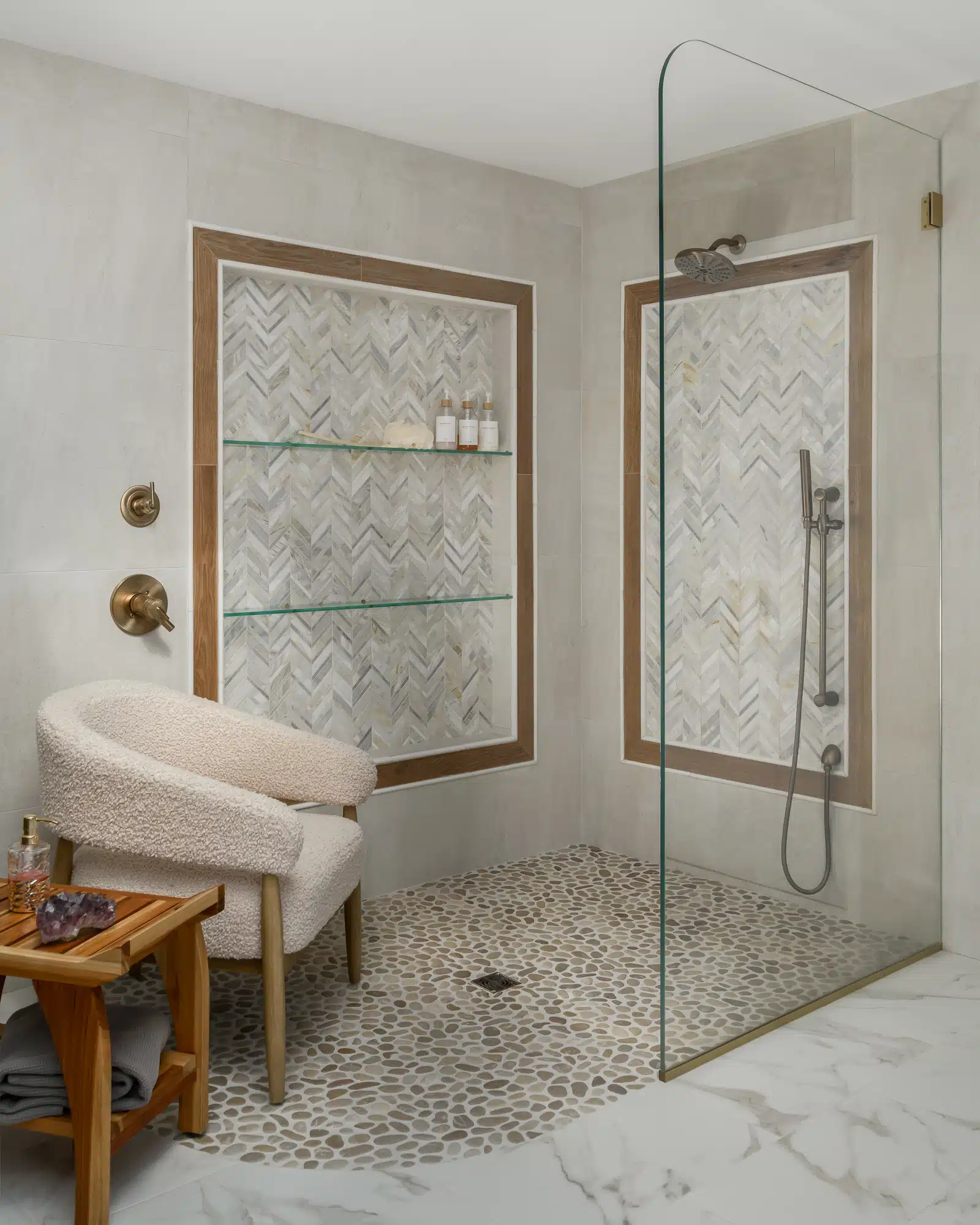 Oversized curbless walk in open shower with 3 levels of glass shelves in large niche, backed by chevron tile rimmed with wood tile planks, pebble shower floor, boucle chair and teak bench, in Medford, NJ.